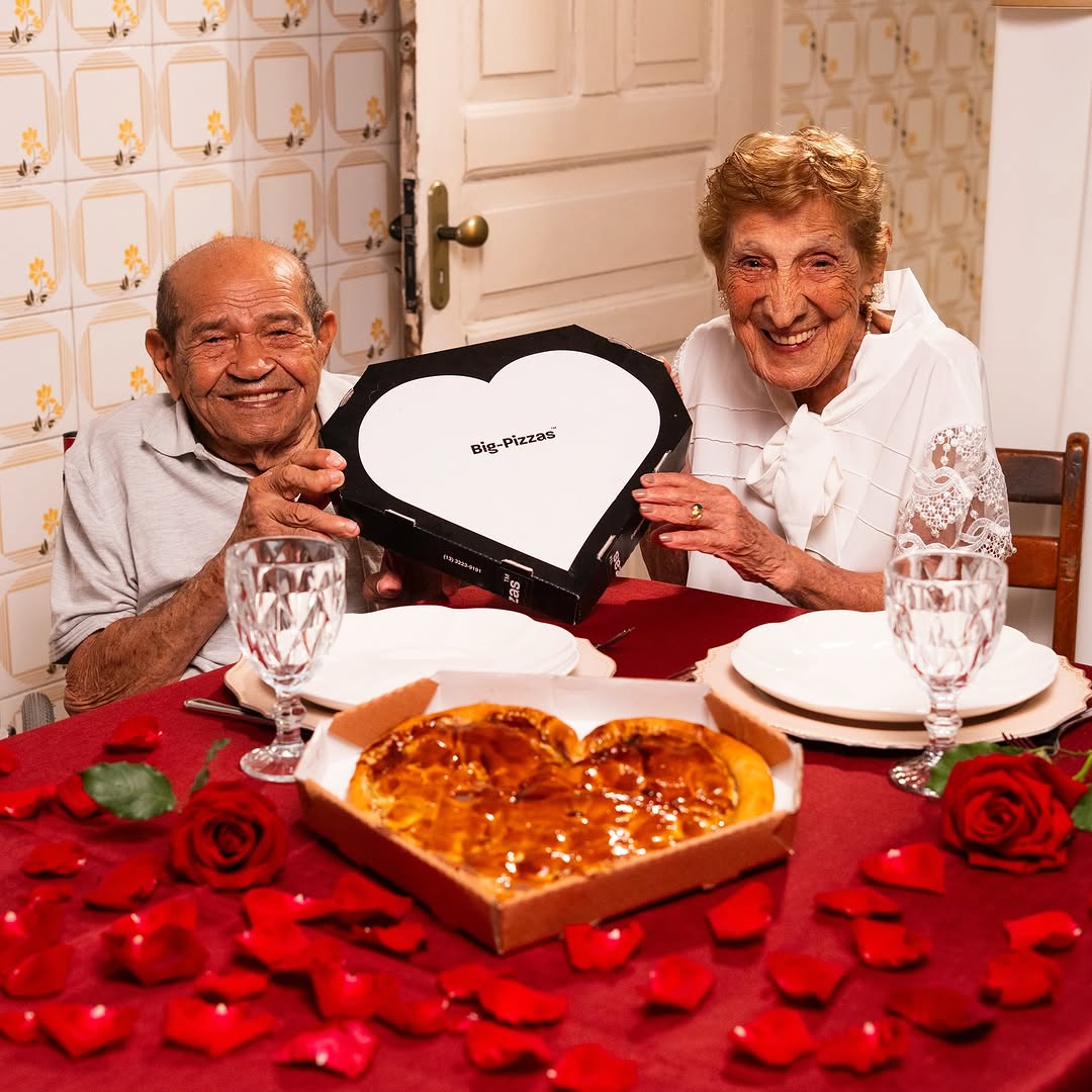 Casal de idosos sorrindo à mesa segurando uma caixa de pizza em formato de coração