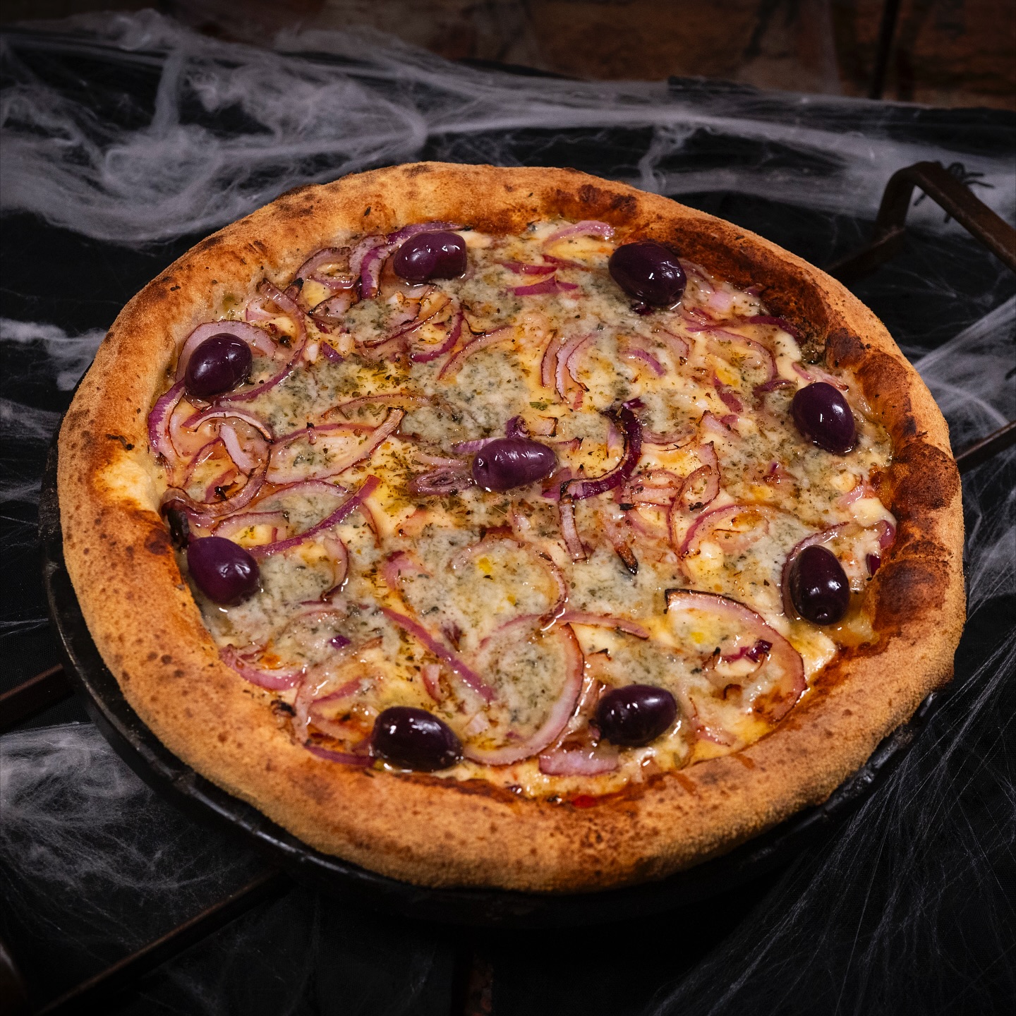 Pizza com cebola roxa, gorgonzola e azeitonas pretas, sobre mesa decorada com teia de aranha