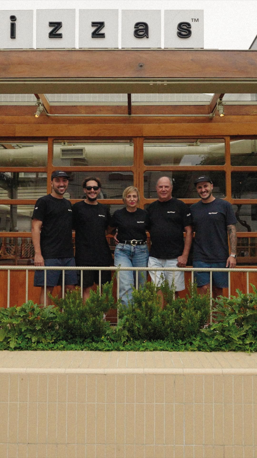 Cinco pessoas da Big de camiseta preta na frente do salão de madeira e vidro, com o letreiro Pizzas acima