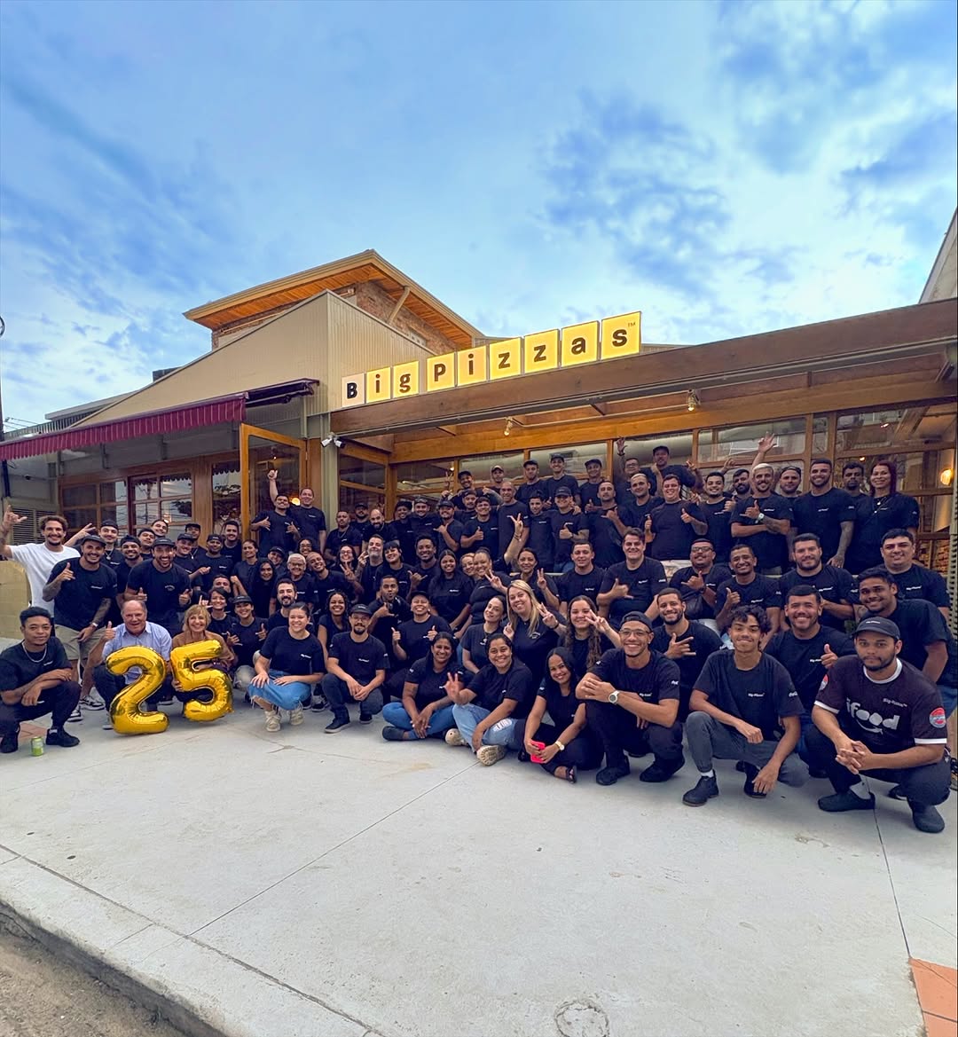 O time inteiro da Big-Pizzas, de camiseta preta, posando na frente da fachada com balões dourados de 25